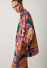 Többszínű blúz merész virágmintával lila, narancs és zöld színben. Magas gallérral és laza fazonnal, sima anyagú, hosszú ujjal rendelkezik.