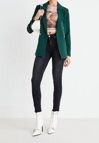 Femme portant un blazer vert croisé, un crop top imprimé, un jean skinny noir, des bottines blanches à talons aiguilles ornées de clous, tenant un sac à main blanc.