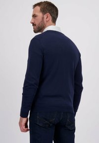 Man met kort haar en een baard, gekleed in een marineblauwe sweater over een wit shirt, gecombineerd met donkerblauwe jeans, met zijn rug naar de camera gericht tegen een lichtgrijze achtergrond.