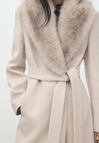Abrigo beige con un amplio y suave cuello de piel sintética, mangas largas y un cinturón a juego en la cintura. Textura lisa y diseño entallado.