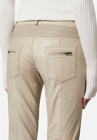 Beige Lederhosen mit einem gerippten Taillenbund und zwei Gesäßtaschen mit Reißverschlüssen. Der Stoff hat eine glatte Textur und ein figurbetontes Design.