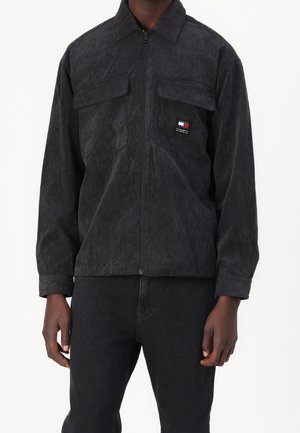 Mann trägt eine dunkelgraue Cord-Reißverschlussjacke mit Klappentaschen auf der Brust und einem kleinen rechteckigen Logo auf der linken Tasche.