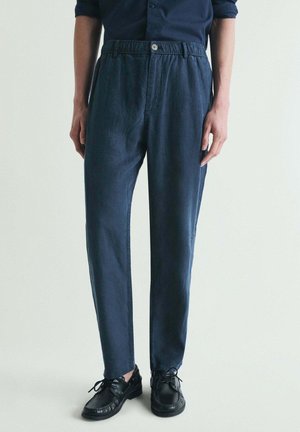 Persona que lleva pantalones casuales azul marino con cintura elástica y zapatos de cuero negros con cordones, de pie frente a un fondo claro y liso.