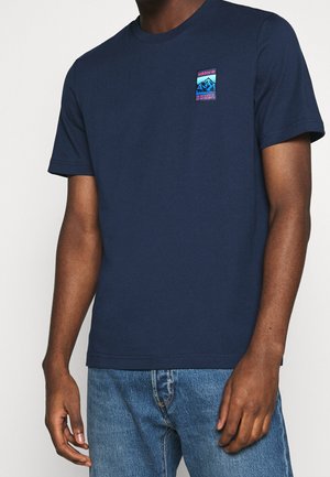 Mężczyzna noszący granatowy t-shirt Adidas z małym kolorowym nadrukiem górskim i współrzędnymi na piersi, w połączeniu z niebieskimi dżinsami.