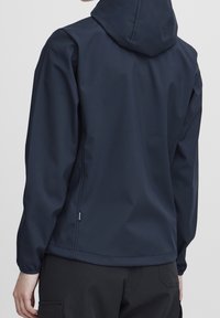 Persona che indossa una giacca a vento blu scuro con cappuccio e pantaloni neri, mostrata di spalle su uno sfondo chiaro e semplice.