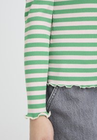 Gestreiftes langärmeliges Oberteil in Grün und Weiß mit Rippstruktur und Rüschensaum an den Bündchen, getragen mit grauen Jeans.