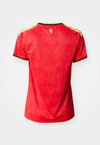 Piros rövid ujjú sportpóló sárga és fekete szegéllyel az ujjakon és a vállakon, finom lángmintákkal és egy kis sárga emblémával a gallér közelében.