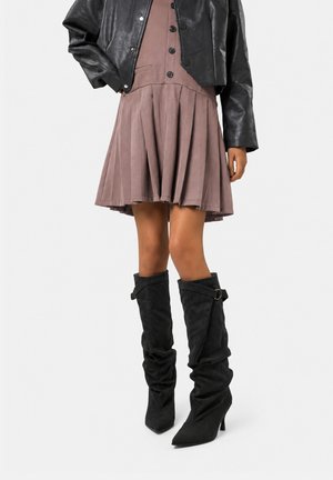 Mujer con un vestido malva plisado, chaqueta de cuero negra corta y botas negras con tacón de punta caída, de pie sobre un fondo blanco.