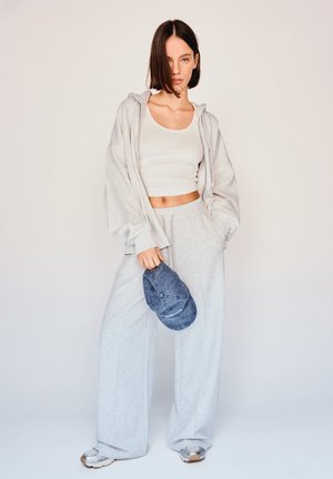 Grå oversized hoodie, matchande sweatpants och en åtsittande vit tanktop. En blå basebollkeps hålls i handen. Mjuka material och avslappnad passform.