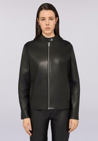 Giacca di pelle nera con colletto alto, zip frontale e maniche lunghe. Texture liscia e design minimalista. Indossata con pantaloni neri.
