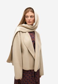 Mujer vestida con un abrigo con capucha beige sobre un vestido estampado en tonos marrón y azul marino, mirando hacia un lado contra un fondo claro.