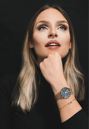 Black watch with a round face and gold mesh strap, worn on a hand resting under the chin. Smooth hair with lighter ends and neutral lips.