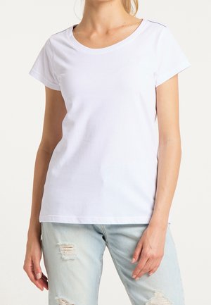 Femme portant un t-shirt blanc uni à manches courtes et un jean bleu clair déchiré, visible du cou jusqu'aux hanches sur un fond neutre.