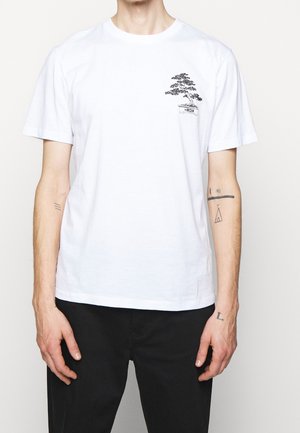 Man wearing white short-sleeve t-shirt with small black bonsai tree design on left chest, paired with black pants.