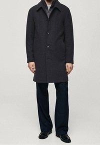 Abrigo de lana gris oscuro con cuello, cierre de botones y bolsillos laterales, combinado con pantalones oscuros y zapatos negros brillantes.