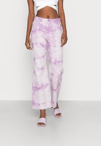 Calças de ganga largas em padrão tie-dye roxo pastel, feitas de denim. Apresentam uma cintura alta e bainha desfiada, combinadas com sandálias de tacão bloco em lilás.