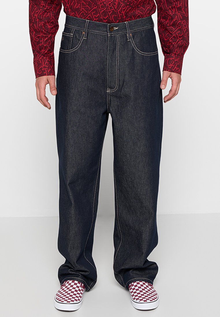 Dunkelblaue Denim-Jeans mit einem lockeren Fit, die kontrastierende weiße Nähte und Fronttaschen aufweisen. Kombiniert mit karierten Slippern.