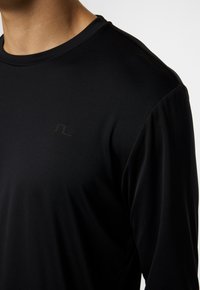 Svart långärmad skjorta gjord av ett mjukt, stretchigt tyg. Har en subtil logo i svart på bröstet. Klassisk rund halsringning.