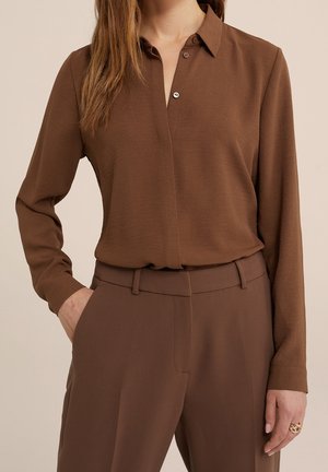 Frau trägt ein braunes, langärmliges Hemd mit Knöpfen, das in passende braune, hoch taillierte Hosen gesteckt ist, mit einer Hand in der Tasche.