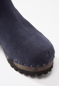 Botte en daim bleu marine à bout arrondi, semelle plateforme en bois, clous métalliques le long de la base et une semelle extérieure noire robuste.