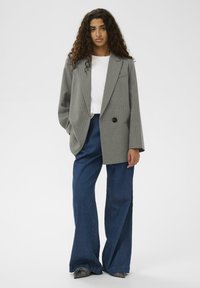 Femeie cu păr creț poartă un sacou oversized din materiale gri în dungi, o cămașă albă, blugi largi albaștri și pantofi ascuțiți de culoare închisă, stând pe un fundal alb.