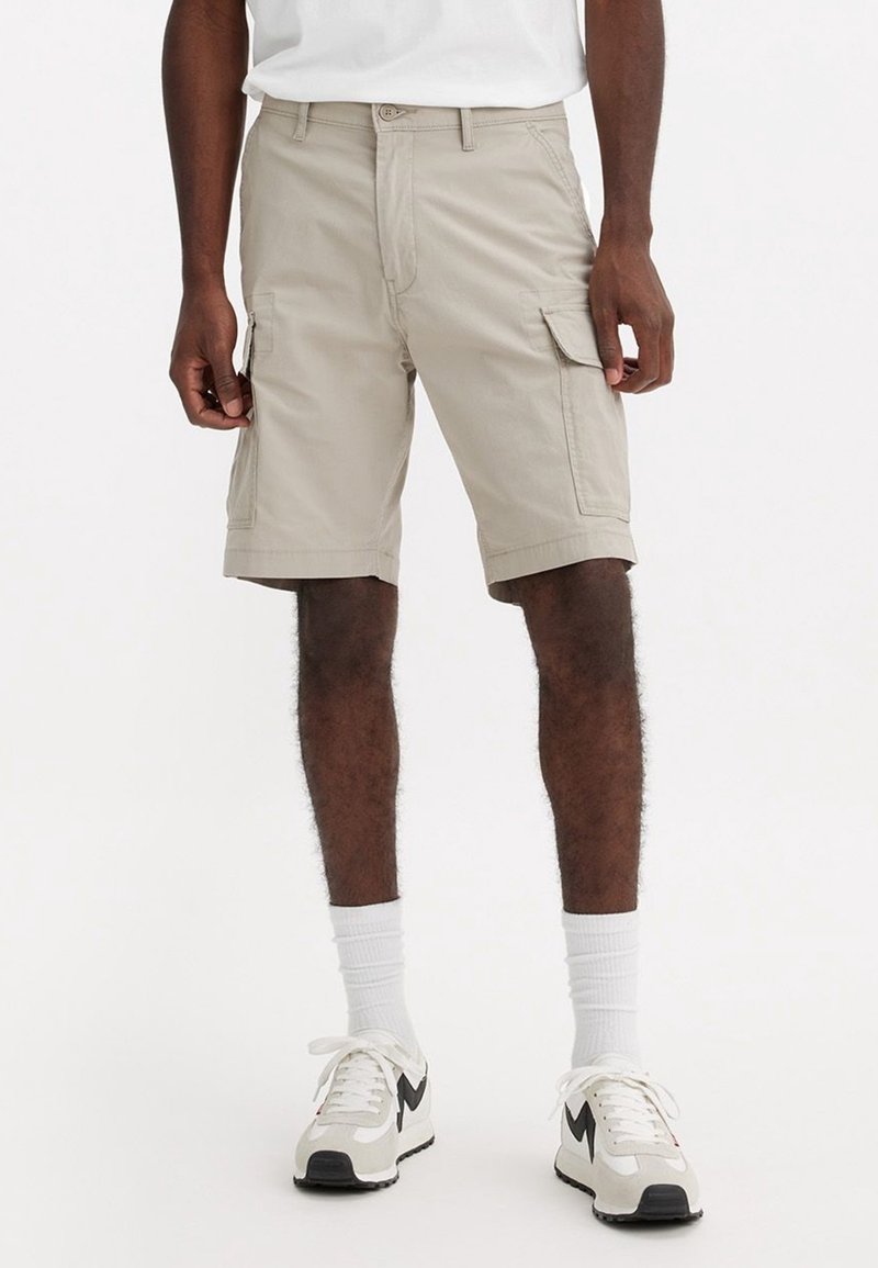 Homme portant un short cargo beige avec des poches latérales, des chaussettes blanches et des baskets blanches avec des touches noires, debout devant un fond uni.