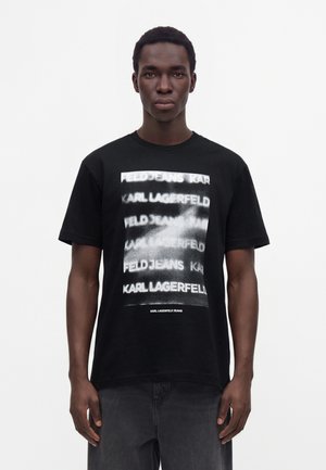 Ung mand iført en sort Karl Lagerfeld jeans t-shirt med hvidt sløret gentaget tekst foran, stående mod en ensfarvet hvid baggrund.
