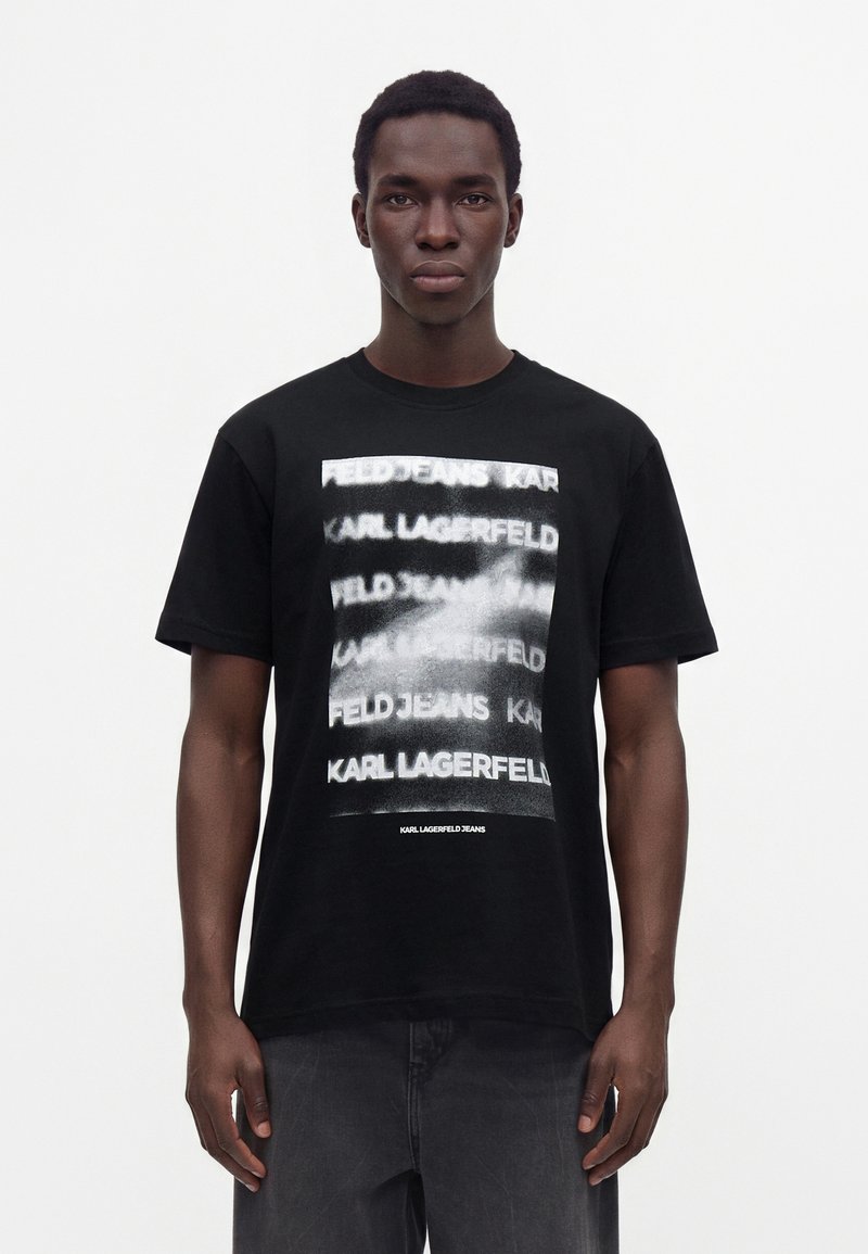 Νεαρός άνδρας που φοράει μαύρο t-shirt Karl Lagerfeld jeans με λευκό, θολό επαναλαμβανόμενο κείμενο μπροστά, στέκεται σε απλό λευκό φόντο.