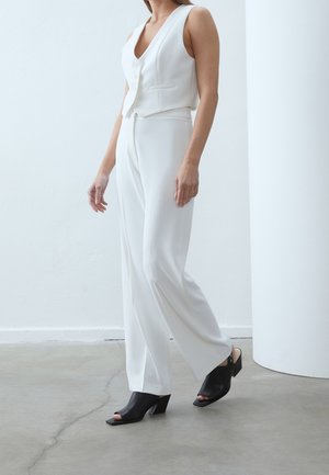 Woman wearing white sleeveless vest and pants paired with black open-toe heeled mules, standing indoors on a concrete floor.