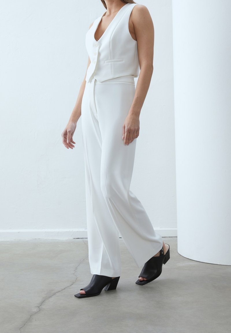 Nainen, joka on pukeutunut valkoiseen hihattomaan paitaan ja housuihin, yhdistettynä mustiin avonaisiin korokepohjaisiin sandaaleihin, seisoo sisätilassa betonilattialla.