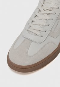 Basket en cuir et daim gris clair avec des lacets blancs, semelle en caoutchouc texturé et bandes d'accent diagonales. Marque sur la languette.