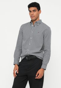 Černobílá košile s kostkovaným vzorem, dlouhými rukávy, zapínací límec a malá logo akcentace; nošena s černými kalhotami a páskem.