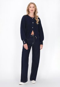 Marineblå strikcardigan med lange ærmer, rund halsudskæring og detaljer med guldknapper. Sammen med højtaljede mørke denimbukser.
