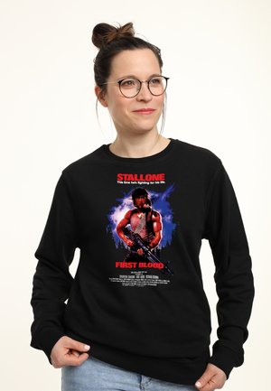 Kvinde med briller og håroppsætning, iført en sort sweatshirt med motiv af en muskuløs soldat, der holder et gevær fra filmen First Blood.