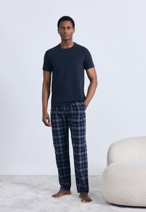 Tricou bleumarin și pantaloni de pijama în carouri albastre, gri și albe. Pantalonii au o croială lejeră cu talie elastică. Barefoot pe un covor deschis la culoare.