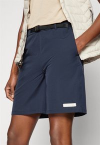 Mørkeblå shorts laget av et lett stoff, med en svart midjeelastikk og en synlig logo-patch på den nedre kanten. Glatt tekstur og skreddersydd passform.