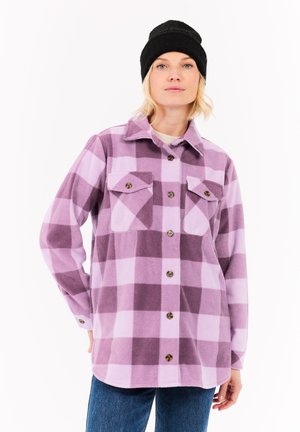 Woman wearing a black knit beanie and oversized purple checkered button-up shirt with two chest pockets, paired with blue jeans.