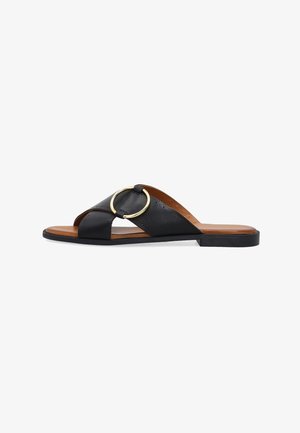 Sort flad slippersandal med krydsede læderremme og et stort rundt spænde i guld, brun indersål og åben tå.