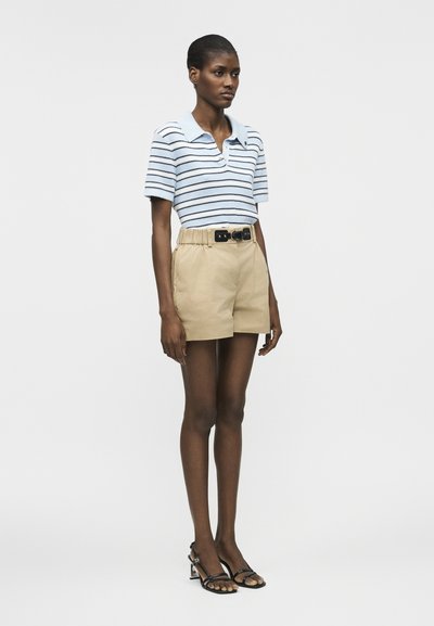 Γυναίκα που φοράει μια ανοιχτογάλαζια και λευκή ριγέ κοντομάνικη μπλούζα πόλο, μπεζ σορτς με ψηλή μέση και μαύρη ζώνη, και μαύρα σανδάλια με τακούνι.