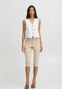Gilet sans manches blanc avec un col en V et cinq boutons dorés, associé à un pantalon beige 7/8 et des sandales à talons noirs.