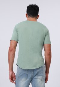 Camiseta de algodón verde claro con dobladillo redondeado y mangas cortas, combinada con jeans de mezclilla azules, vista desde la parte de atrás.