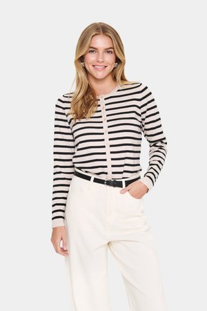 Long-sleeve, striped top in beige and black, featuring a round neckline and button closure. Paired with high-waisted, white trousers.