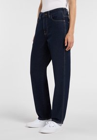 Person som bär mörkblå raka jeans med kontrastsömmar och vita snörskor, ståendes mot en enfärgad bakgrund.