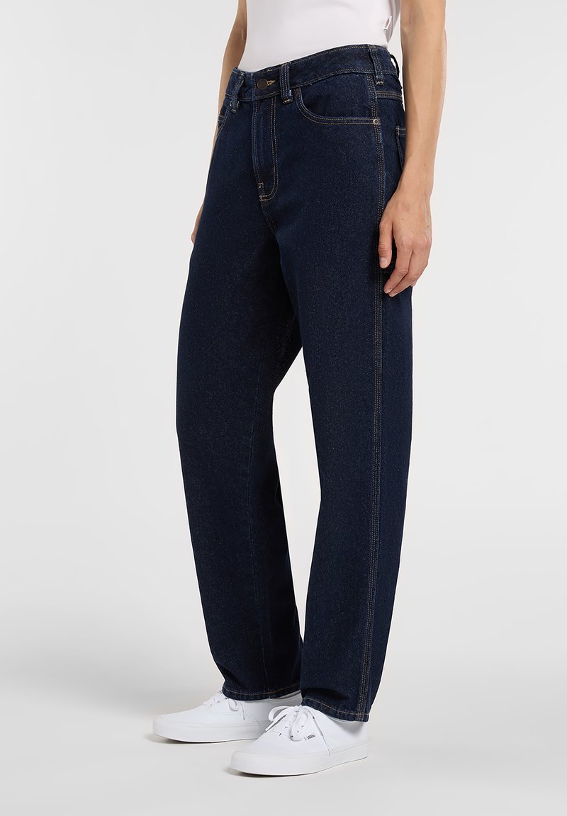 Person wearing dark blue straight-leg jeans with contrast stitching and white lace-up sneakers, standing against a plain background.