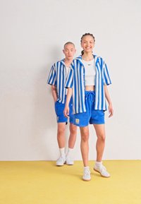 Blauwe en witte gestreepte shirts met korte mouwen en korte broeken van zachte stof, gecombineerd met witte sneakers. Eenvoudige achtergronden benadrukken de outfits.