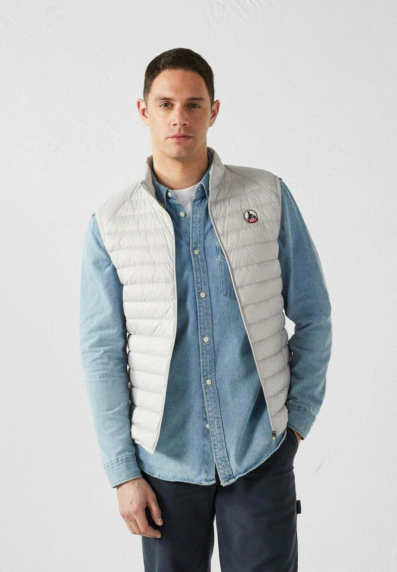 Gilet trapuntato grigio chiaro con colletto alto, indossato sopra una camicia di denim. Presenta un logo rotondo sul lato sinistro del petto e una superficie liscia.