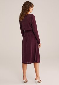 Rochie lungă burgund cu mâneci lungi, cu talie ajustată și fustă fluidă; realizată dintr-un material lucios, cu o textură moale și un luciu subtil.