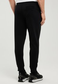 Mees, kes kannab musti kitsenevaid pükse ja musti tenniseid, seisab seljaga meile, taustal hele ja ühetaoline pind.