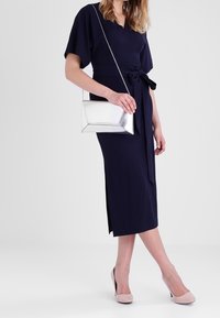 Navy midi dress with short sleeves, belted waist, and side slit. Paired with a silver rectangular handbag and nude heels.