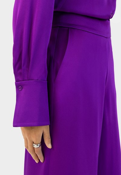 Paarse zijde blouse met een knoopmanchet, met een soepele textuur en vloeiende ontwerp, deels zichtbaar met een hand die op de stof rust.